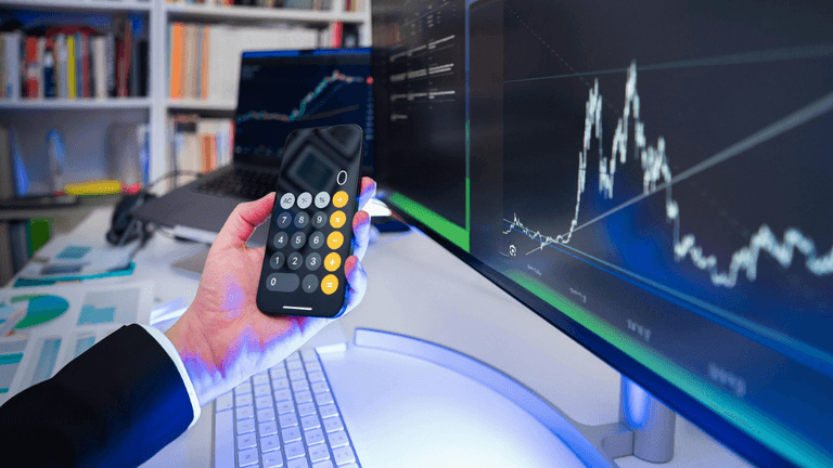 Hand holding a calculator with financial charts displayed on a large monitor