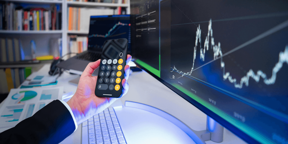 Hand holding a calculator with financial charts displayed on a large monitor