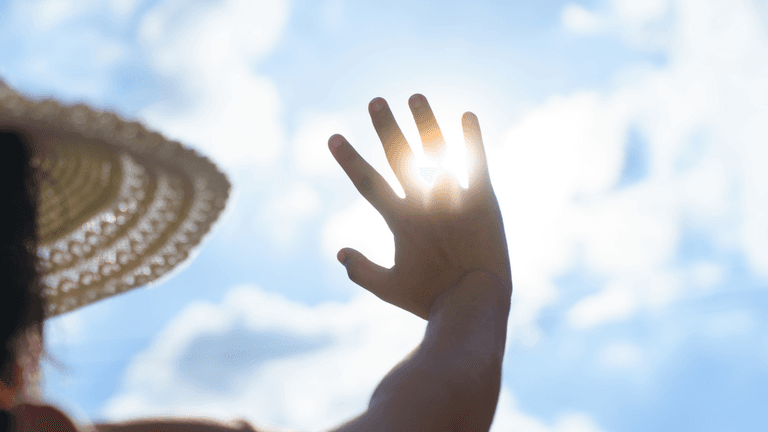 A person in a straw hat raises their hand toward the sun in a cloudy sky