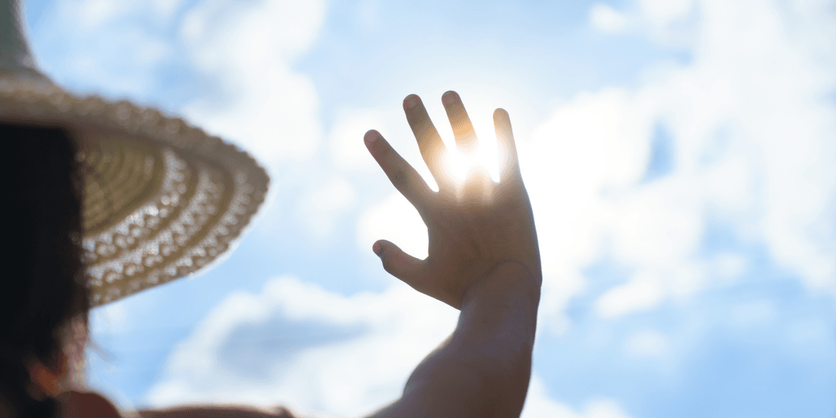 A person in a straw hat raises their hand toward the sun in a cloudy sky