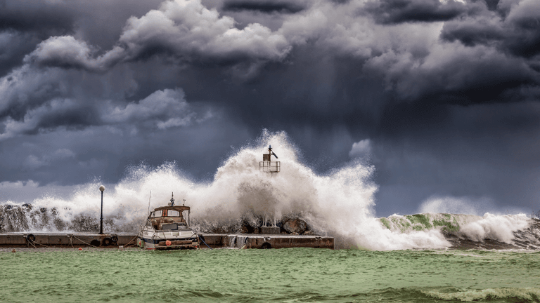 Waves crash against a pier with an anchored boat under stormy skies