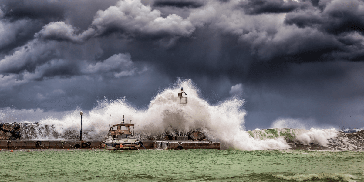 Waves crash against a pier with an anchored boat under stormy skies